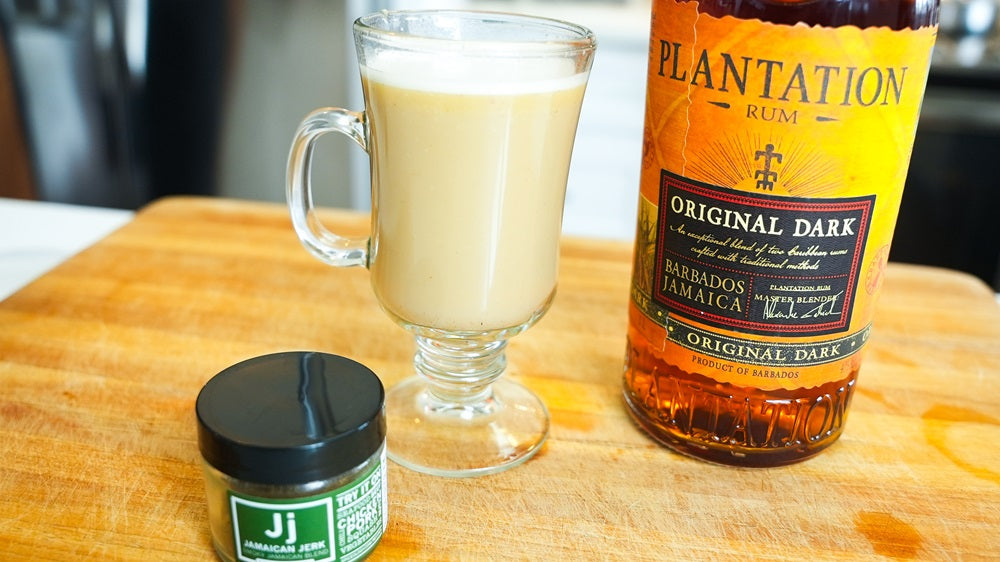 Glass of Jamaican Jerk Hot Buttered Rum Cocktail next to a bottle of rum and jar of Spiceology Jamaican Jerk blend