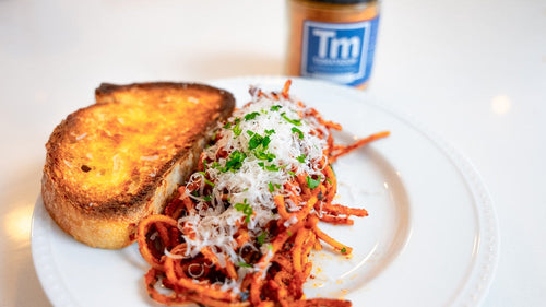 Plated Assassin's Spaghetti topped with grated Parmesan and parsley with a slice of garlic bread on the plate next to a jar of Spiceology Tomatador blend