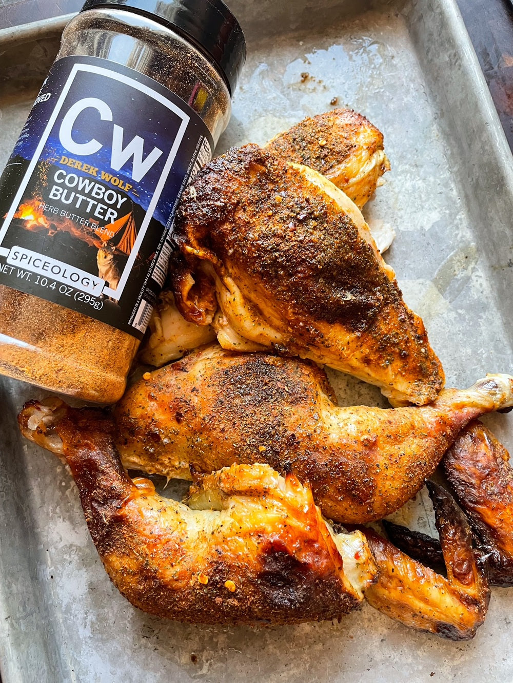 Quartered whole chicken on a sheet pan next to a Spiceology X Derek Wolf Cowboy Butter seasoning bottle