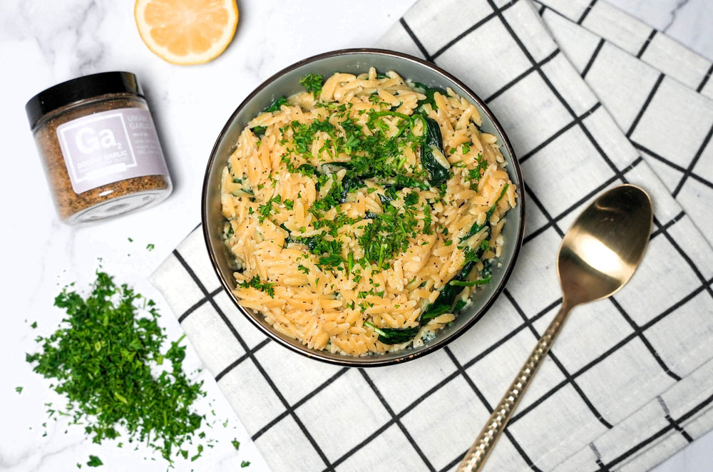 Bowl of Spinach Orzo next to a serving spoon, chopped parsley, a lemon wedge, and a jar of Spiceology Double Garlic seasoning