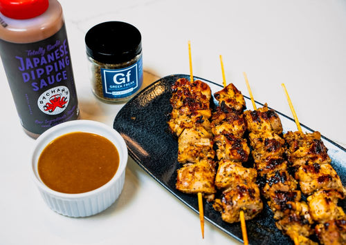 Plated grilled chicken thighs on skewers next to a bowl and bottle of Bachan's Totally Garlic Dipping Sauce  and jar of Spiceology Greek Freak