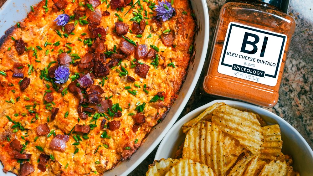 Baking dish of Bleu Cheese Buffalo Chicken Dip next to a bowl of potato chips and a jar of Spiceology Bleu Cheese Buffalo seasoning