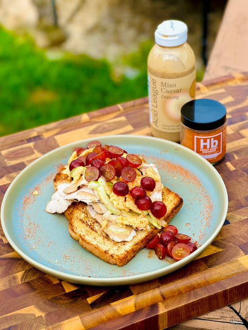 Chicken Salad Tartine with Marinated Grapes next to a bottle of Acid League Miso Caesar and jar of Spiceology Smoky Honey Habanero 