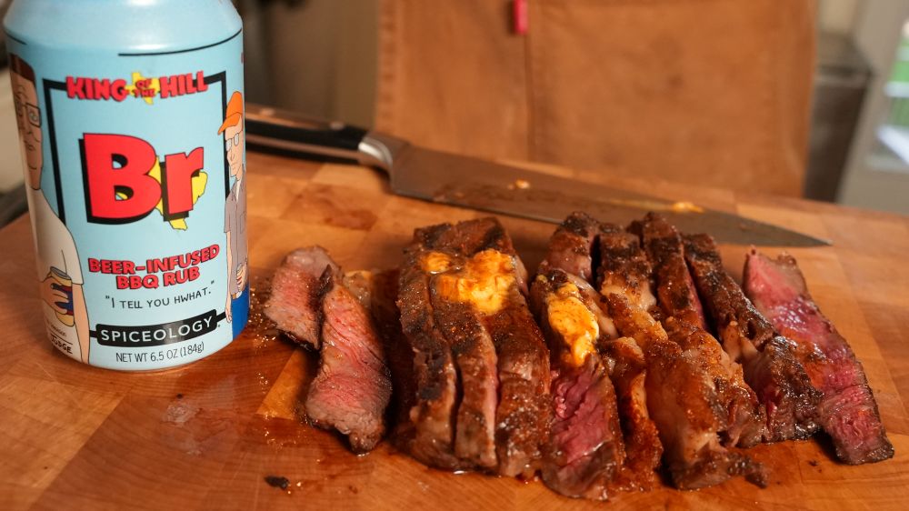 Sliced steak topped with compound butter on a wood cutting board next to a Spiceology X King of the Hill Beer-Infused BBQ Rub beer can