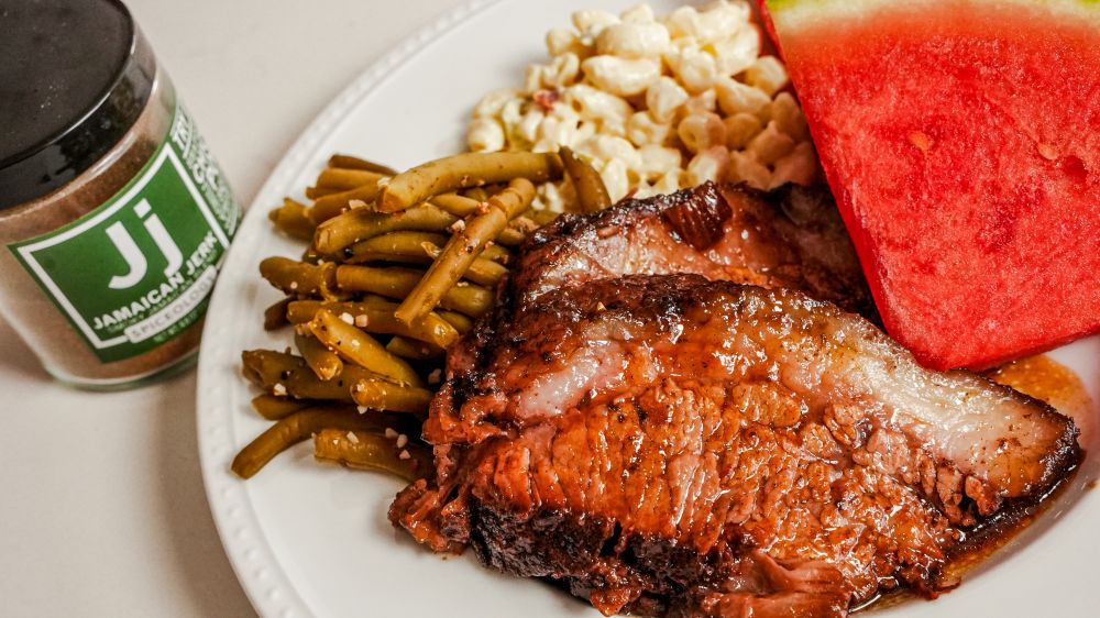 Plated Crockpot Brisket with green beans, macaroni salad, and sliced watermelon next to a jar of Spiceology Jamaican Jerk seasoning
