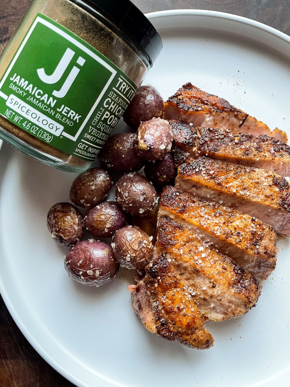 Plated slices of pork chop with roasted mini potatoes next to a jar of Spiceology Jamaican Jerk seasoning.