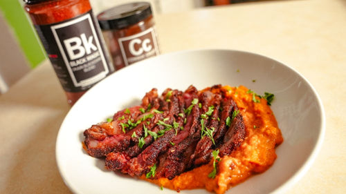 Plated sliced steak and sweet potato mash next to jars of Spiceology Black Magic and Cowboy Crust