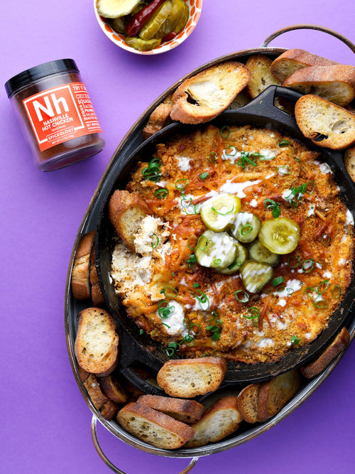 A tray of nashville hot chicken dip with bread and pickles on a purple background