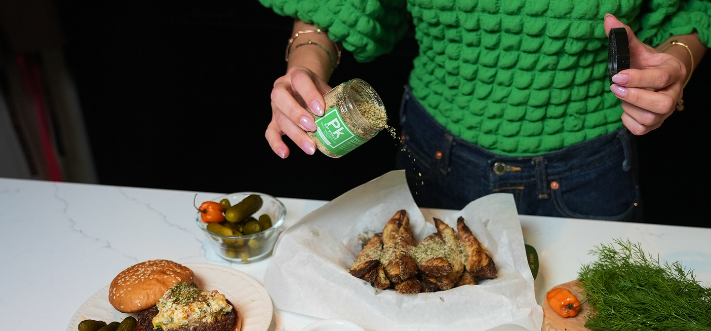 Person in green sweater holding a jar of Spiceology Hot Pickle with food on a table