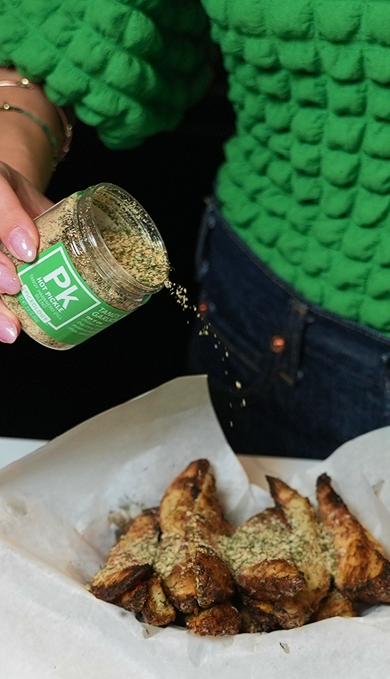 Person sprinkling a green spice mix from a jar labeled 'PK' onto fried food on a white paper.