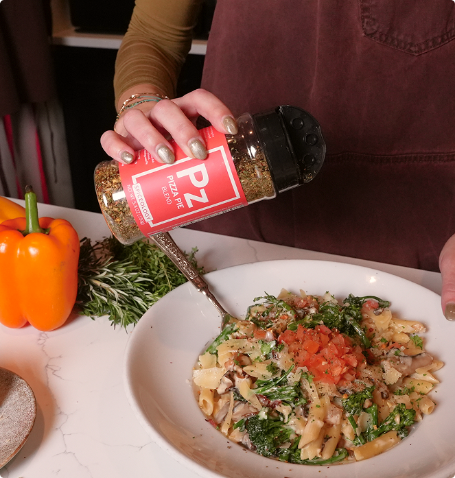 Person seasoning pasta dish with Spiceology Pizza Pie seasoning bottle, surrounded by fresh vegetables.