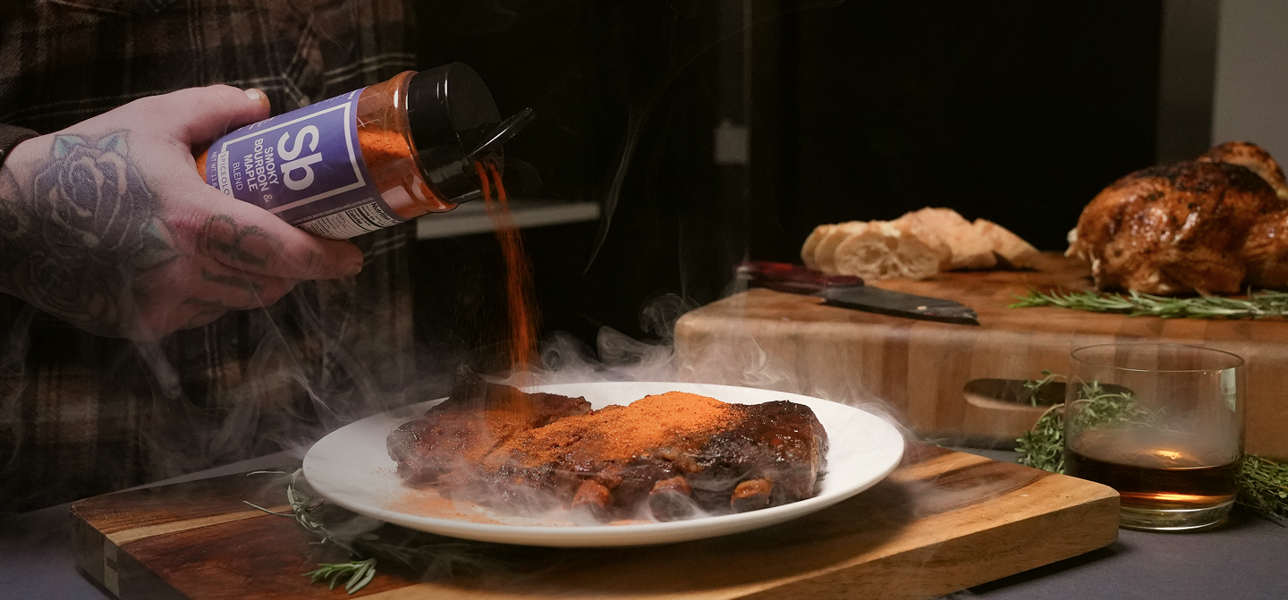 Person seasoning ribs with a bottle of Spiceology Smokey Bourbon & Maple blend on a wooden cutting board.