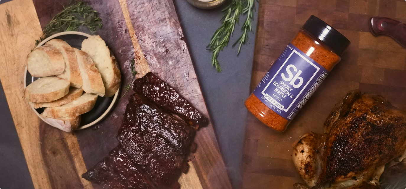 Wooden cutting board with roasted meat, bread, and a bottle of Spiceology Smokey Bourbon & Maple blend on a wooden surface.