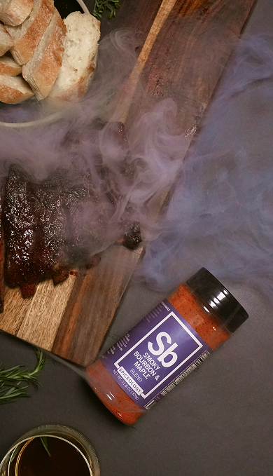 Spice bottle labeled Spiceology Smokey Bourbon & Maple blend with smoking ribs and bread on a wooden board