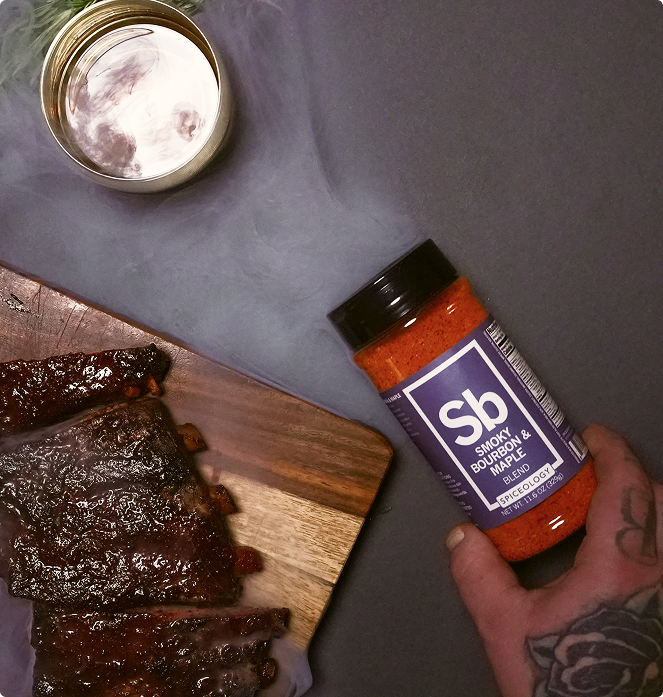 Hand holding a jar of Spiceology Smokey Bourbon & Maple blend next to smoked meat on a wooden board.