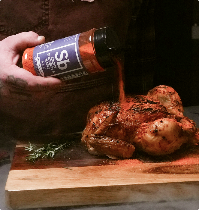Person pouring seasoning from a bottle labeled Spiceology Smokey Bourbon & Maple blend onto a roasted chicken on a wooden cutting board.