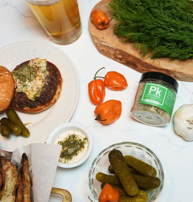 Gourmet food platter with a burger, pickles, peppers, and a jar of Spiceology Hot Pickle Tangy Habanero Dill Blend on a white surface.