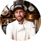 Circular cropped headshot of a man in a white chef coat in a professional kitchen