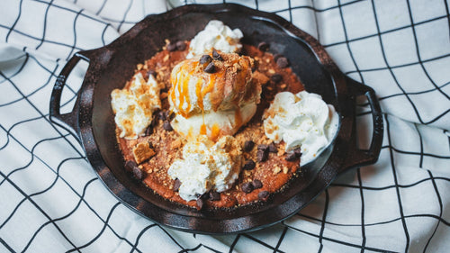 Cast iron skillet with a baked dip or dessert topped with whipped cream on a plaid cloth