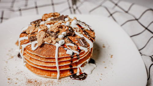 Stack ofS'mores Pancakes featuring  Fluffy pancakes seasoned with Spiceology's S’mores blend, stacked high and finished with toasted marshmallows, Hershey’s chocolate chunks, graham cracker crumbles, and Spiceology's Carbonated Sugar for an experiential dining moment on a white plate.