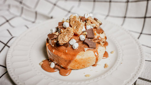 S'mores Cinnamon Roll featuring a warm, gooey cinnamon roll layered with Spiceology's S’mores seasoning, finished with toasted marshmallow fluff, melted chocolate, and a dusting of graham cracker crumble on a white plate with a checkered cloth background