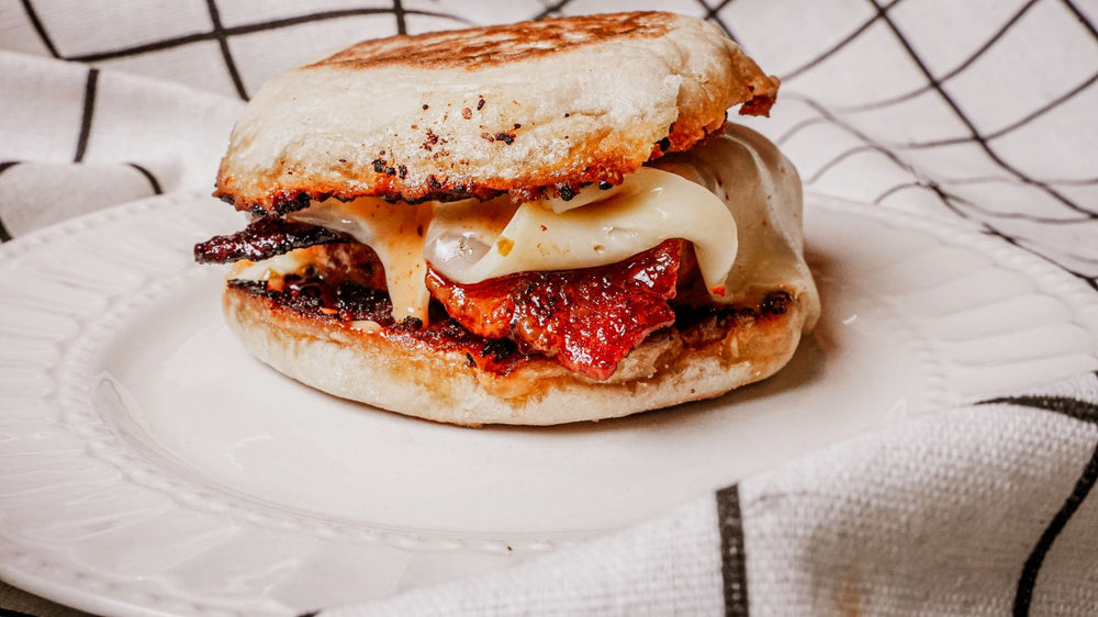 Candied Bacon Breakfast Sandwich featuring a toasted English muffin stacked with fluffy egg, melty cheese, and Spiceology Smoky Honey Habanero–glazed bacon for the perfect balance of sweet heat and savory comfort on a white plate with a checkered cloth background