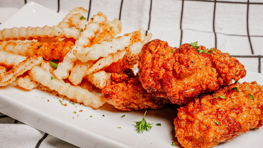 Nashville Hot Chicken Tenders featuring Crispy fried tenders tossed in Spiceology's Nashville Hot seasoning for balanced sweet heat, finished with an optional Smoky Honey Habanero hot honey drizzle next to a pile of seasoned fries on a white plate with a checkered tablecloth.