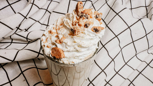 Brown Butter Cookie Shake featuring Creamy vanilla custard spun with Spiceology's Brown Butter Cookie seasoning and loaded with cookie crumbles, finished with a swirl of whipped cream in a glass on a checkered fabric background