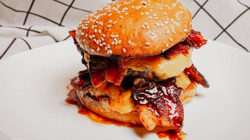 Candied Baconator featuring double beef patties, American cheese, and signature sauces, topped with candied bacon made with Spiceology's Smoky Honey Habanero seasoning for a bold swicy twist. The sweet heat caramelization amplifies the sandwich’s signature smoky richness while keeping the indulgent, bacon-forward identity fans expect. A high-impact flavor upgrade that taps into swicy momentum without disrupting a proven classic on a white plate with a checkered cloth background
