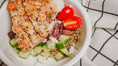 Greek Freak Herb Chicken Bowl featuring Grilled chicken breast tossed in Spiceology's Greek Freak seasoning post-cook for bright herbaceous flavor, served over warm brown rice with cucumber, tomato, olives, feta, and an olive oil finish, then garnished with crispy shallots on a checkered tablecloth