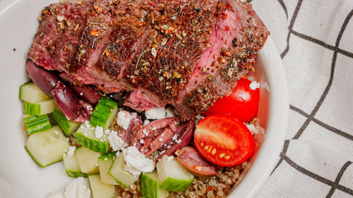 Greek Freak Beef Bowl featuring Steak seasoned with Spiceology's Greek Freak seasoning, served over warm quinoa with crisp cucumbers, tomatoes, olives, and fresh greens, finished with a final sprinkle of Greek Freak for bright, herbaceous flavor with a checkered cloth background