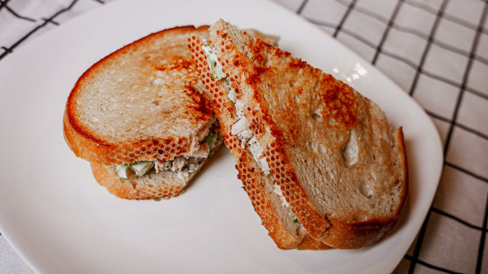 Greek Freak Chicken Salad Sandwich is a classic favorite with a Mediterranean twist. Tender, shredded chicken is grilled to perfection and tossed in our bold Greek Freak seasoning. Packed with vibrant herbs, citrusy notes, and savory spices on a white plate with a checkered tablecloth in the background