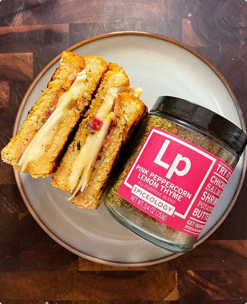 Pink Peppercorn Lemon Thyme Bacon, Brie & Apple Grilled Cheese sandwich cut in half on a plate with a jar of Spiceology Pink Peppercorn Lemon Thyme seasoning.