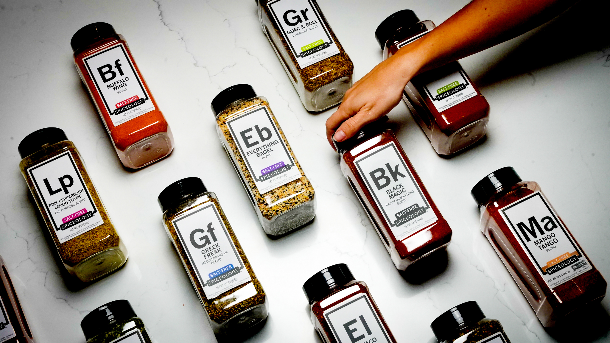 Assorted spice jars with labels on a white surface, one jar is being picked up.