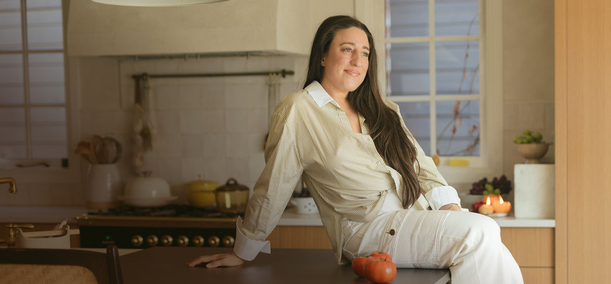 A woman with long dark hair sits on a kitchen island, wearing a cream-colored button-up shirt and matching pants. The kitchen has light wood cabinets, a tiled backsplash, and warm ambient lighting.
