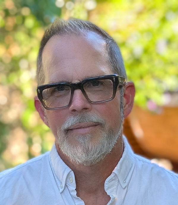 Outdoor portrait of a man wearing glasses in casual attire with green foliage in the background