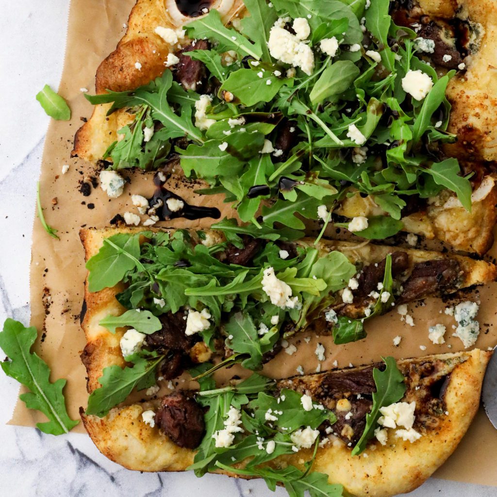 a steak pizza with bleu cheese and arugula on a white background