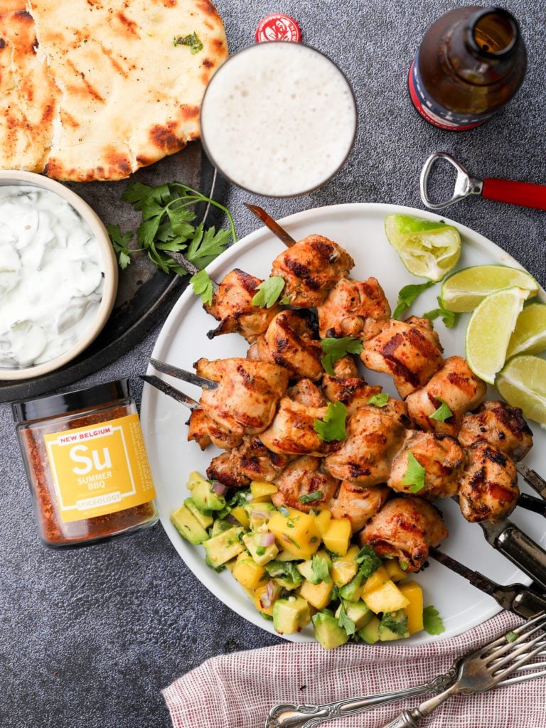 a plate of chicken skewers with a jar of spice, a beer, naan bread and tzatziki sauce