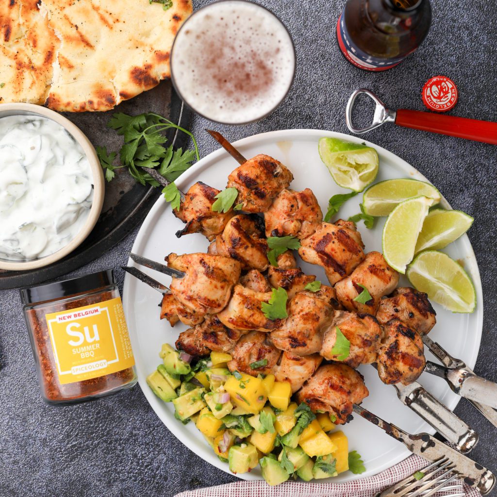 chicken on skewers with lime, avocado mango relish, tzatziki, naan bread, beer and a jar of spices
