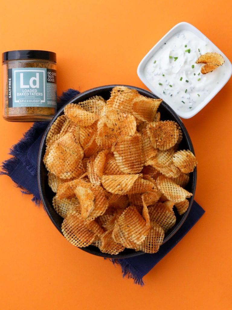 a bowl of waffle potato chips on an orange background with a bowl of sour cream dip and a jar of spices